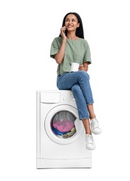 Beautiful woman with cup of drink sitting on washing machine and talking on phone against white background