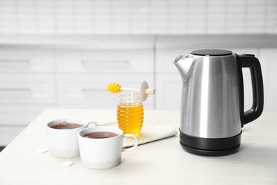Photo of Modern electric kettle, cups of tea and honey on wooden table in kitchen