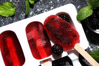 Photo of Mold with tasty blackberry ice pops on dark textured table, flat lay. Fruit popsicle