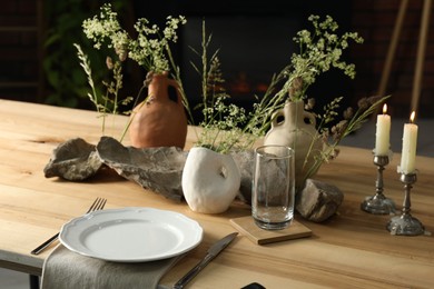 Photo of Set of clean dishware, vases with beautiful flowers, stones and burning candles on wooden table in stylish dining room