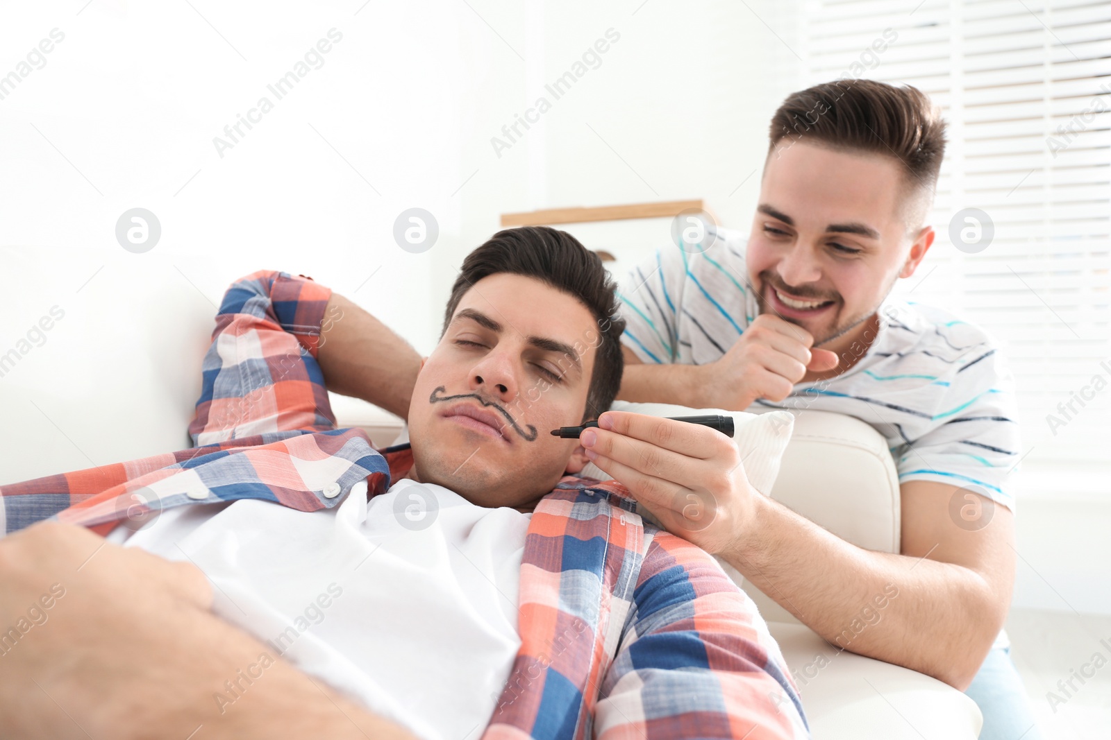 Photo of Young man drawing mustache on face of sleeping friend indoors. April fool's day