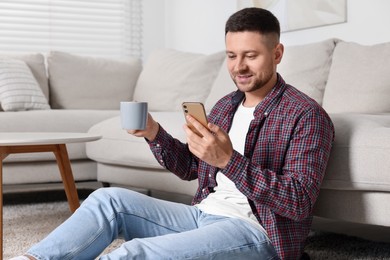 Photo of Handsome man with cup of drink sending message via smartphone at home