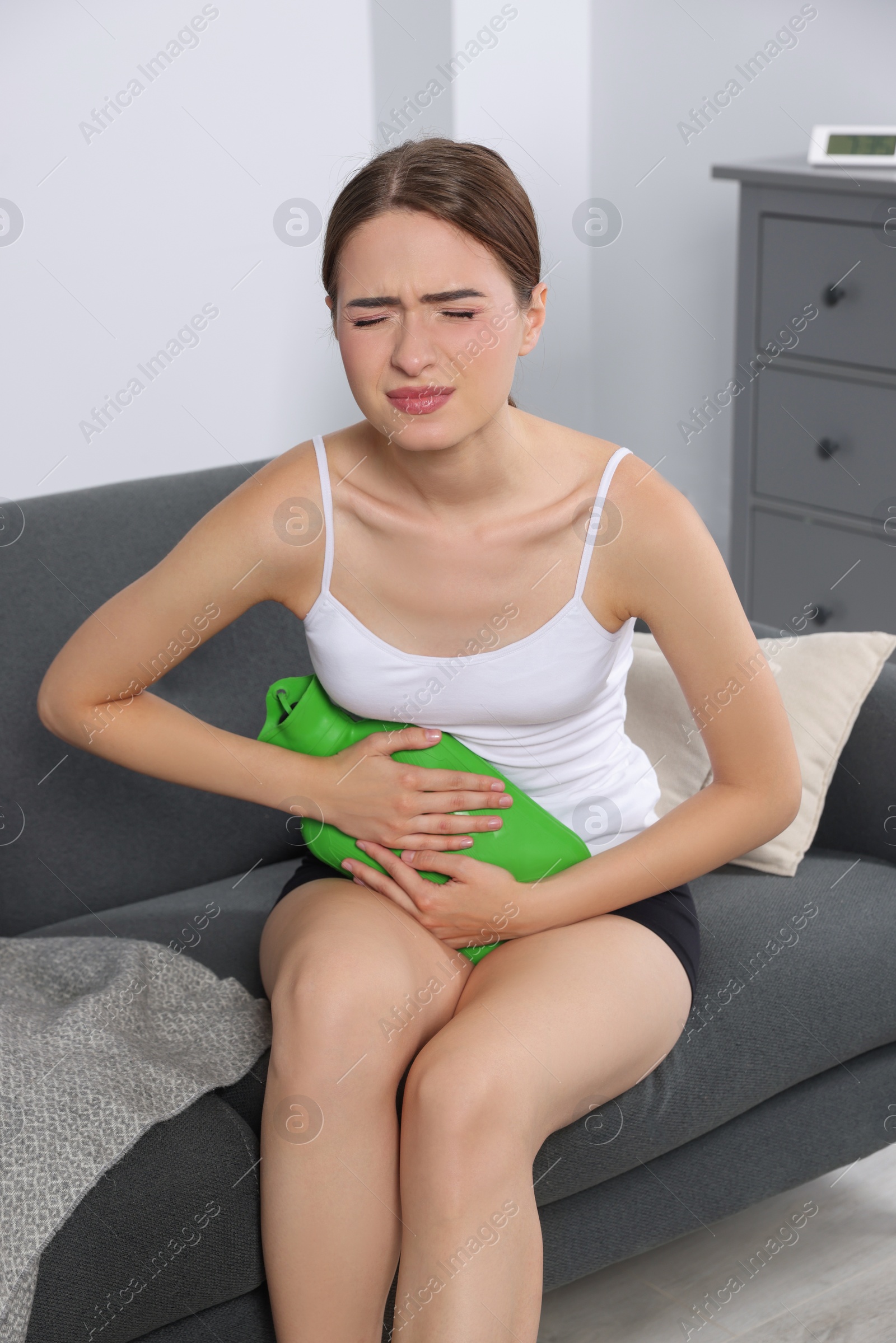 Photo of Young woman using hot water bottle to relieve cystitis pain on sofa at home