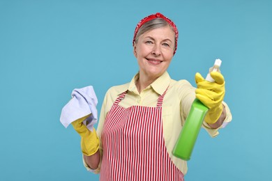 Happy housewife with spray bottle and rag on light blue background