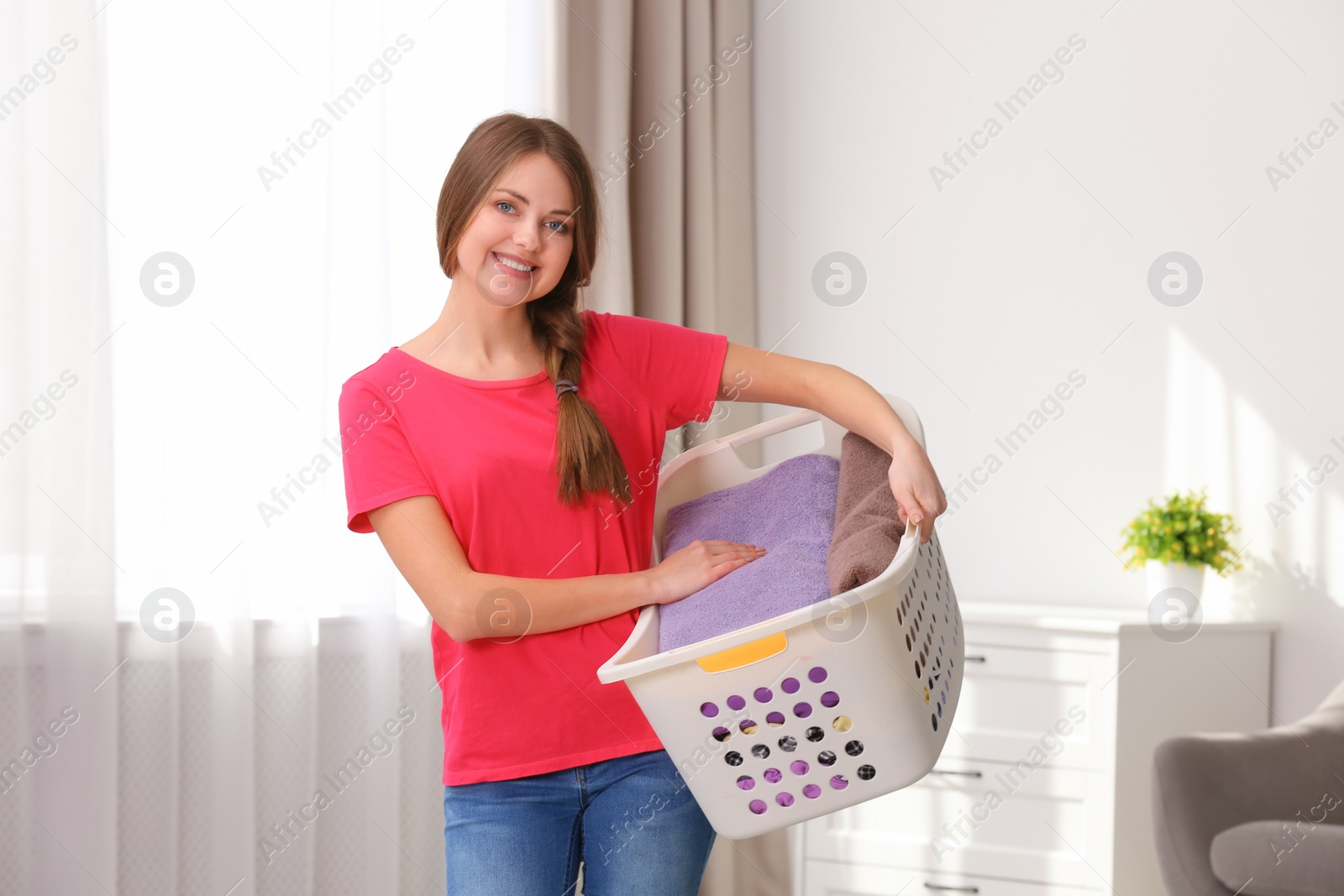 Photo of Happy young woman holding basket with laundry at home