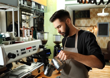 Barista pouring milk into metal pitcher near coffee machine at bar