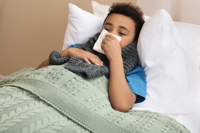 African-American boy with scarf and tissue blowing nose in bed indoors. Cold symptoms
