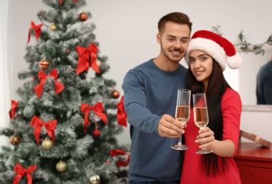 Photo of Young beautiful couple with glasses of champagne near Christmas tree at home