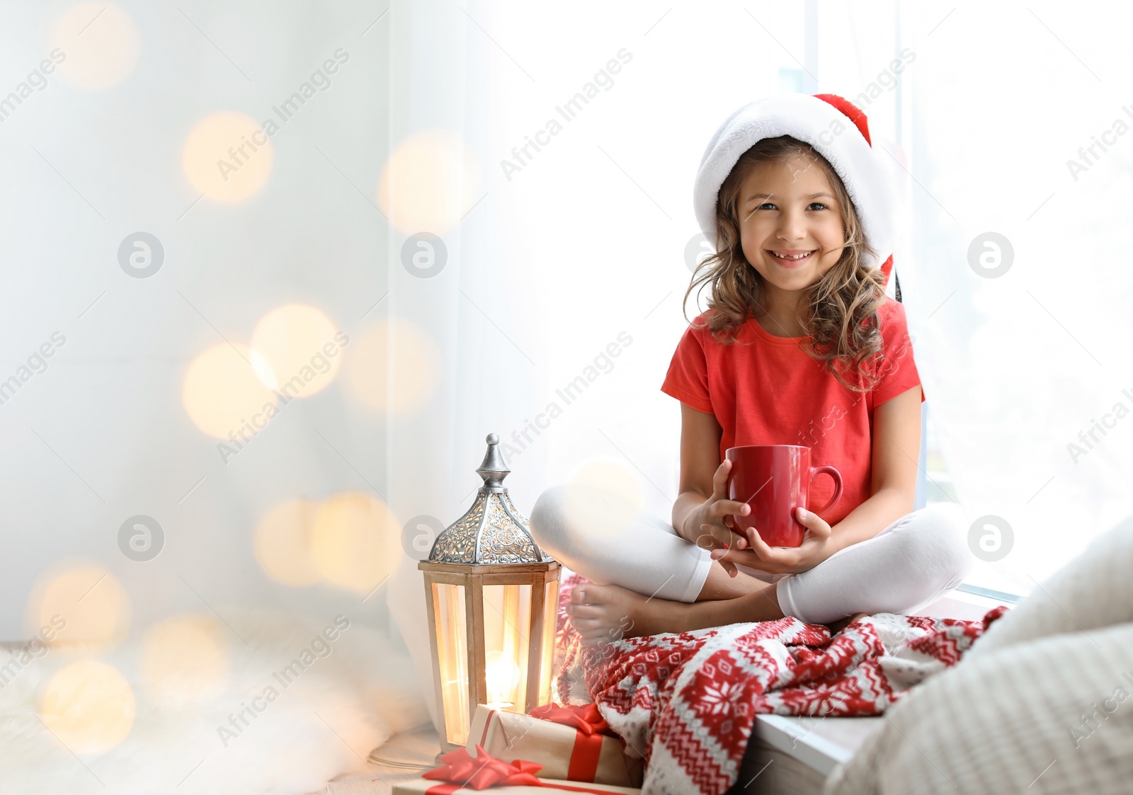 Photo of Cute little child in Santa hat with cup of cocoa sitting on windowsill at home. Christmas celebration