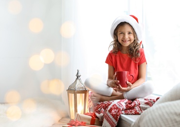 Cute little child in Santa hat with cup of cocoa sitting on windowsill at home. Christmas celebration