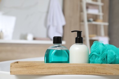 Wooden bath tray with bottles of shower gels and mesh pouf on tub indoors