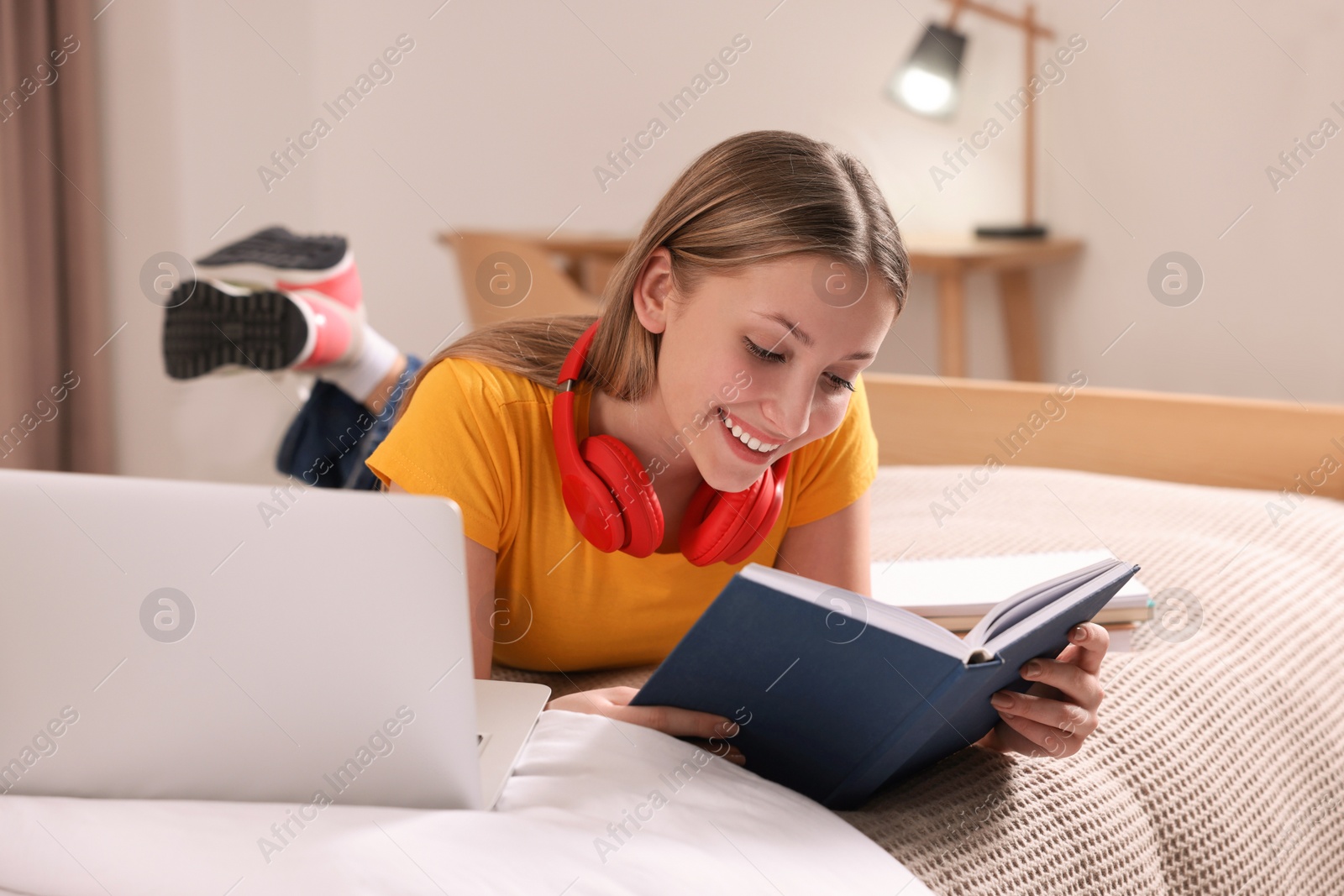 Photo of Online learning. Teenage girl reading book near laptop on bed at home