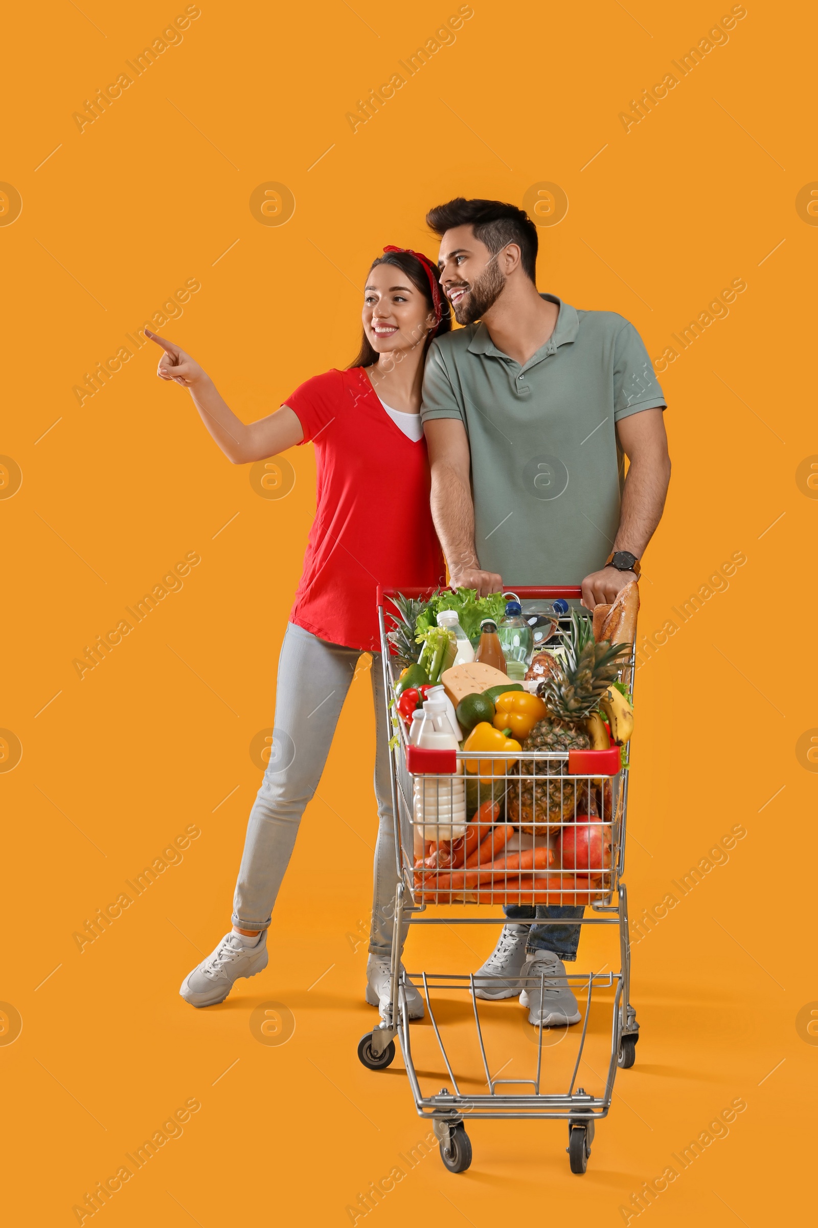 Photo of Young couple with shopping cart full of groceries on yellow background