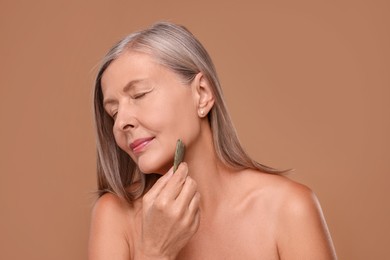 Woman massaging her face with jade gua sha tool on brown background