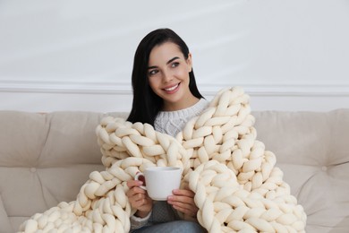 Photo of Young woman with chunky knit blanket and cup on sofa at home