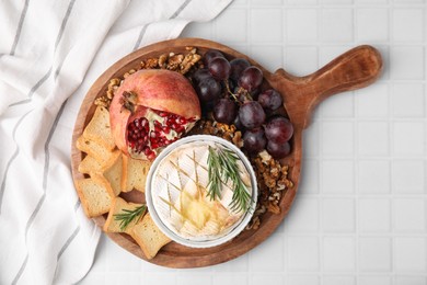 Photo of Board with tasty baked camembert, croutons, grapes, walnuts and pomegranate on white tiled table, top view