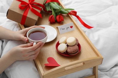 Photo of Tasty breakfast served in bed. Woman with tea, macarons, gift box, flowers and I Love You card at home, closeup