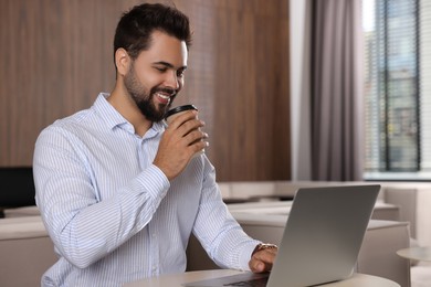 Photo of Happy young man with paper cup of coffee working on laptop at table in office