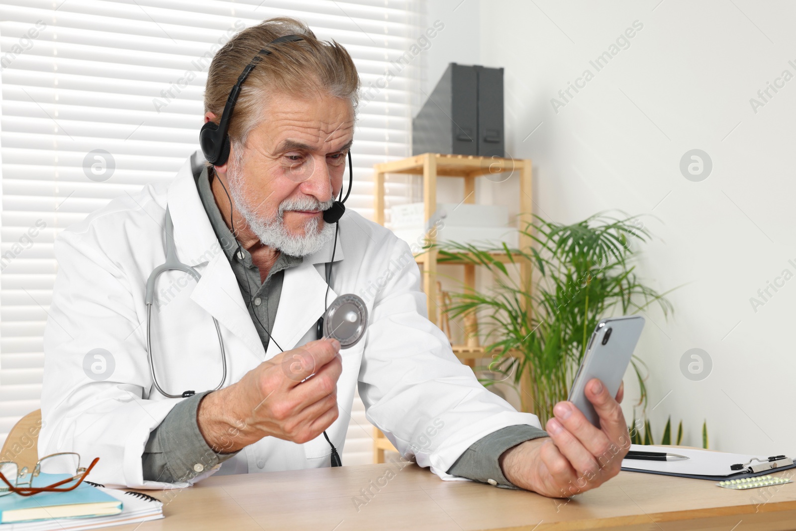 Photo of Senior doctor with phone consulting patient at wooden desk in clinic. Online medicine