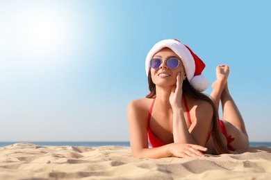 Beautiful young woman in Santa hat and bikini on beach, space for text. Christmas vacation