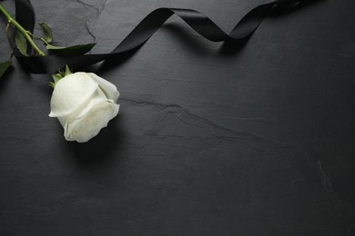 Photo of White rose and ribbon on black table, flat lay with space for text. Funeral symbols