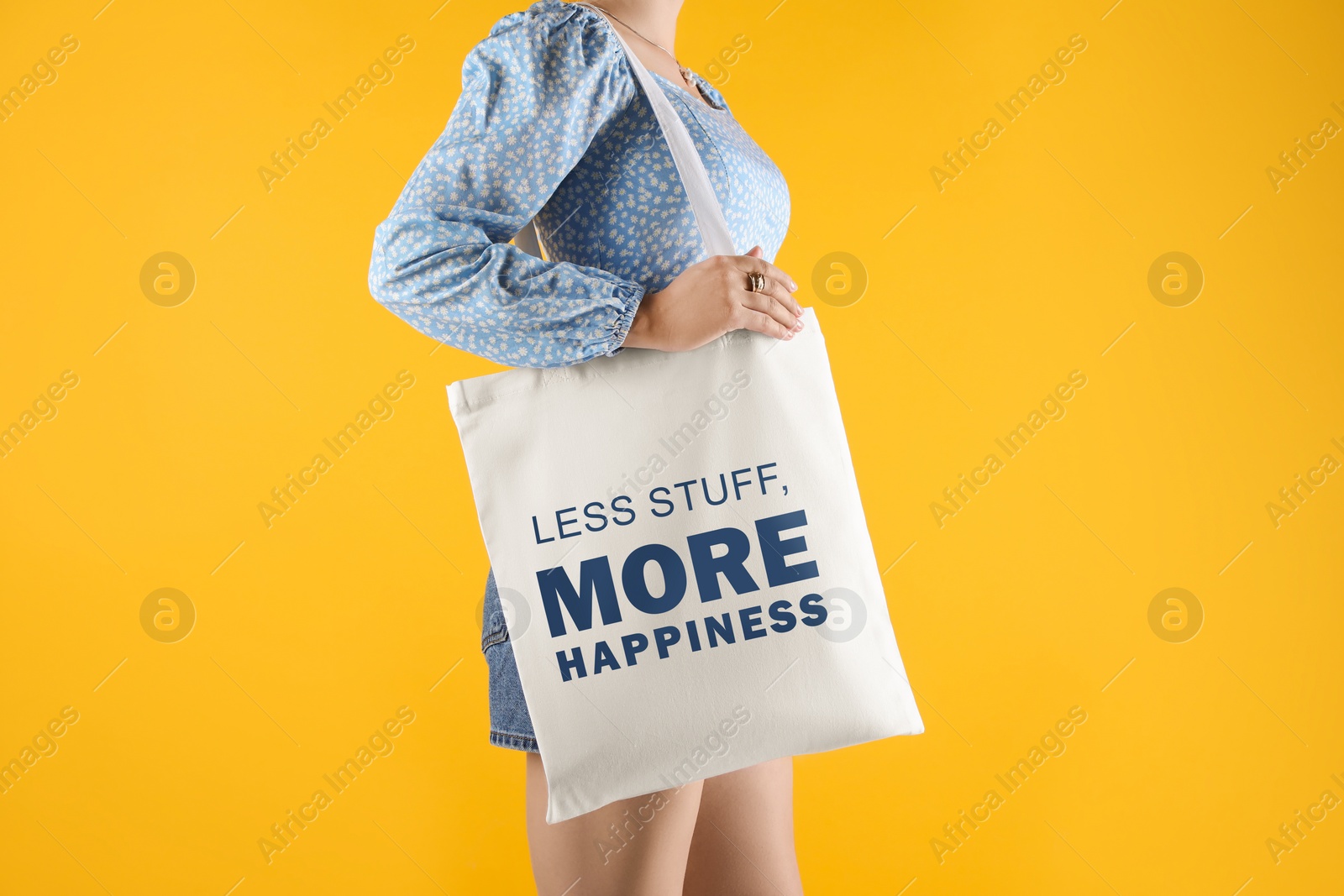 Image of Motivational vibes. Woman holding textile bag with phrase Less Stuff More Happiness on yellow background, closeup