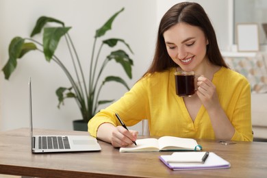 Photo of Smiling woman with cup of coffee writing in notebook at wooden table indoors