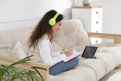 Photo of African American woman with notebook and headphones studying on sofa at home. Distance learning