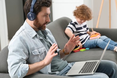 Photo of Father working remotely on laptop while his son playing with toys at home. Man having video chat with colleagues