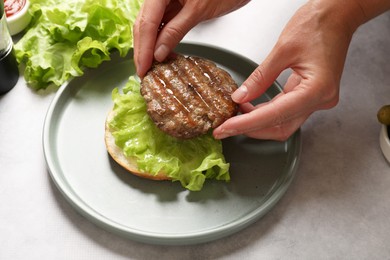 Photo of Woman making tasty hamburger with fried patty, lettuce and bun at light gray table, closeup