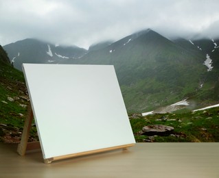 Image of Wooden easel with blank canvas on table and beautiful mountain landscape. Space for text