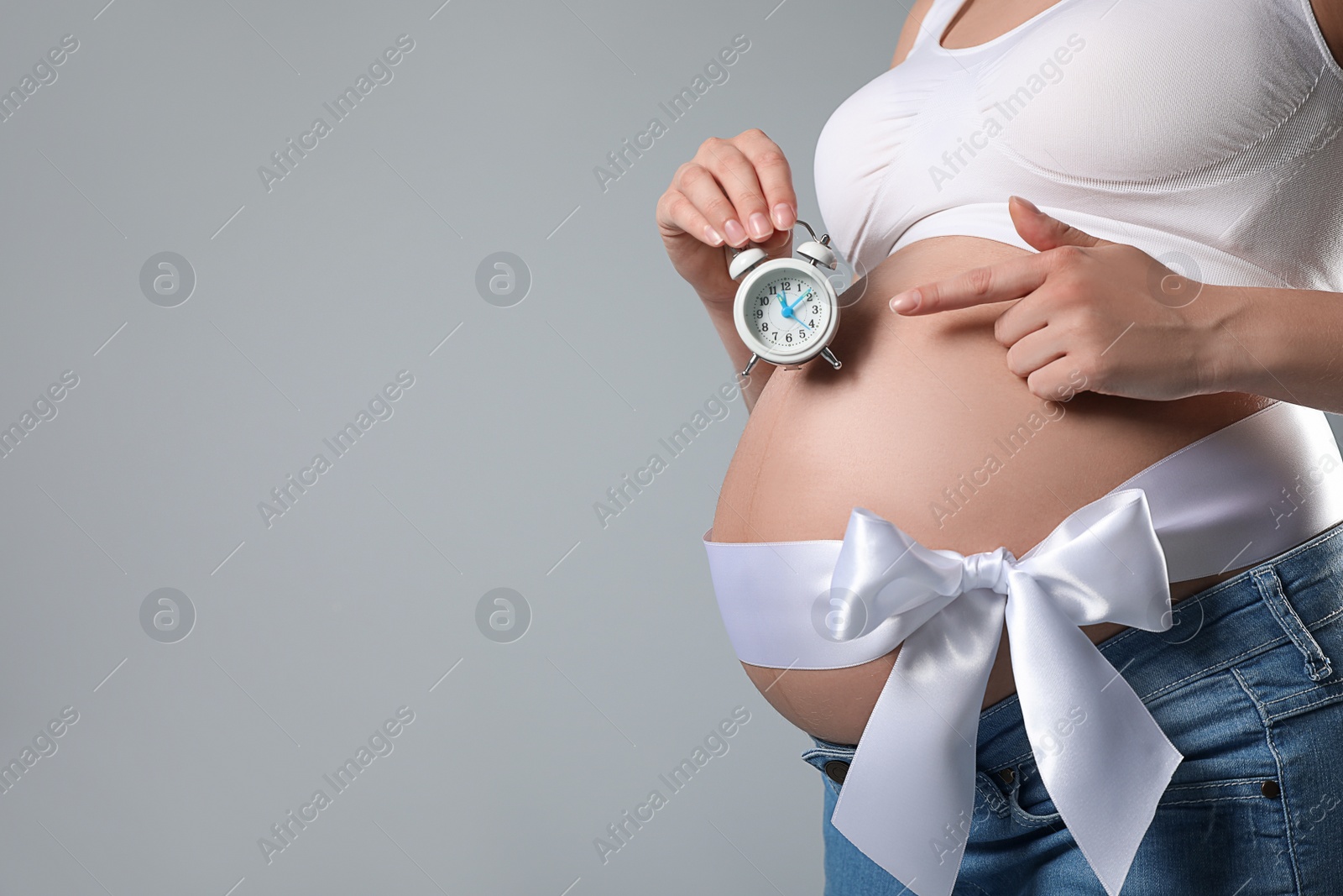 Photo of Young pregnant woman with alarm clock and bow on grey background, closeup. Time to give birth