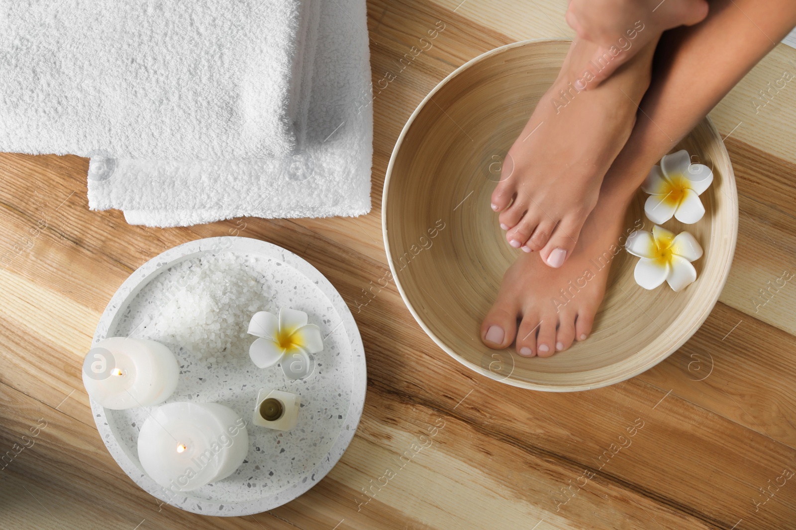 Photo of Woman soaking her feet in dish with water and flowers on wooden floor, top view. Spa treatment