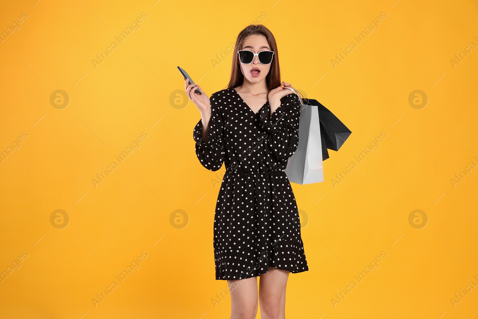 Photo of Surprised young woman with shopping bags and smartphone on yellow background. Big sale