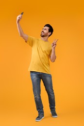 Smiling man taking selfie with smartphone and showing peace sign on yellow background