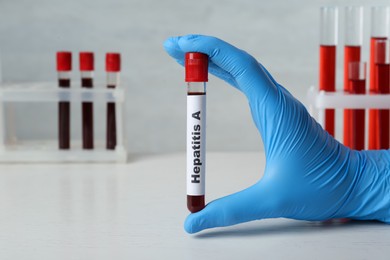 Scientist holding tube with blood sample and label Hepatitis A at white wooden table, closeup