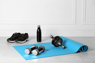Photo of Exercise mat, dumbbells, ab roller, shoes and bottle of water on light wooden floor indoors
