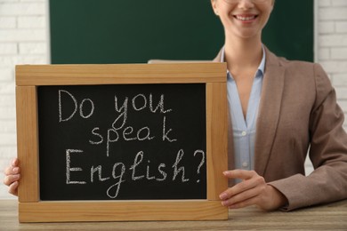 Photo of Teacher holding small chalkboard with inscription Do You Speak English? at table in classroom, closeup