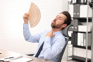 Photo of Bearded businessman waving hand fan to cool himself at table in office