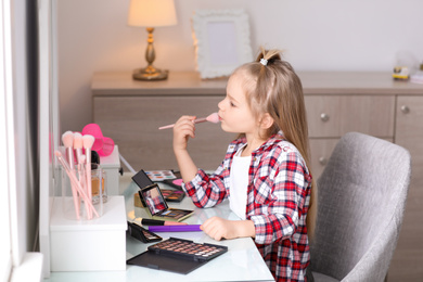 Adorable little girl applying makeup at dressing table indoors
