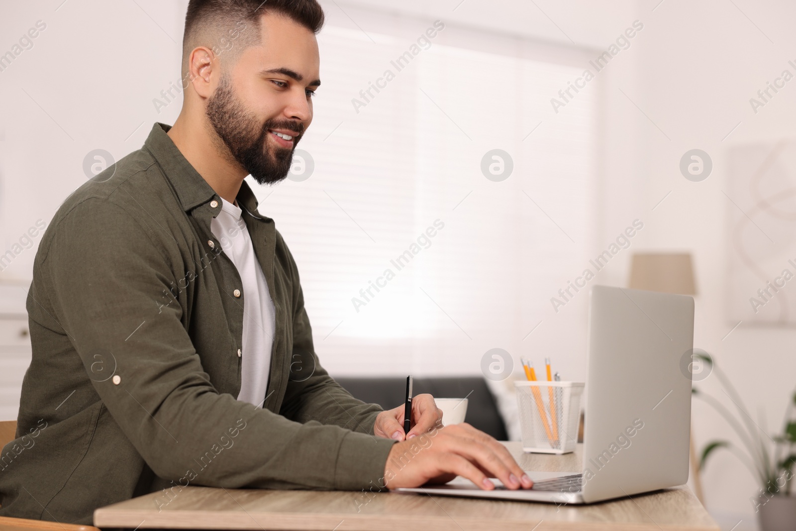 Photo of Young man writing in notebook while working on laptop at wooden table indoors