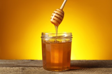 Pouring honey from dipper into jar at wooden table against golden background