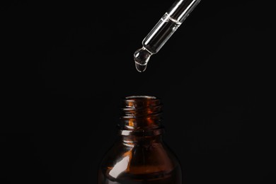 Dripping cosmetic serum from pipette into bottle on black background, closeup