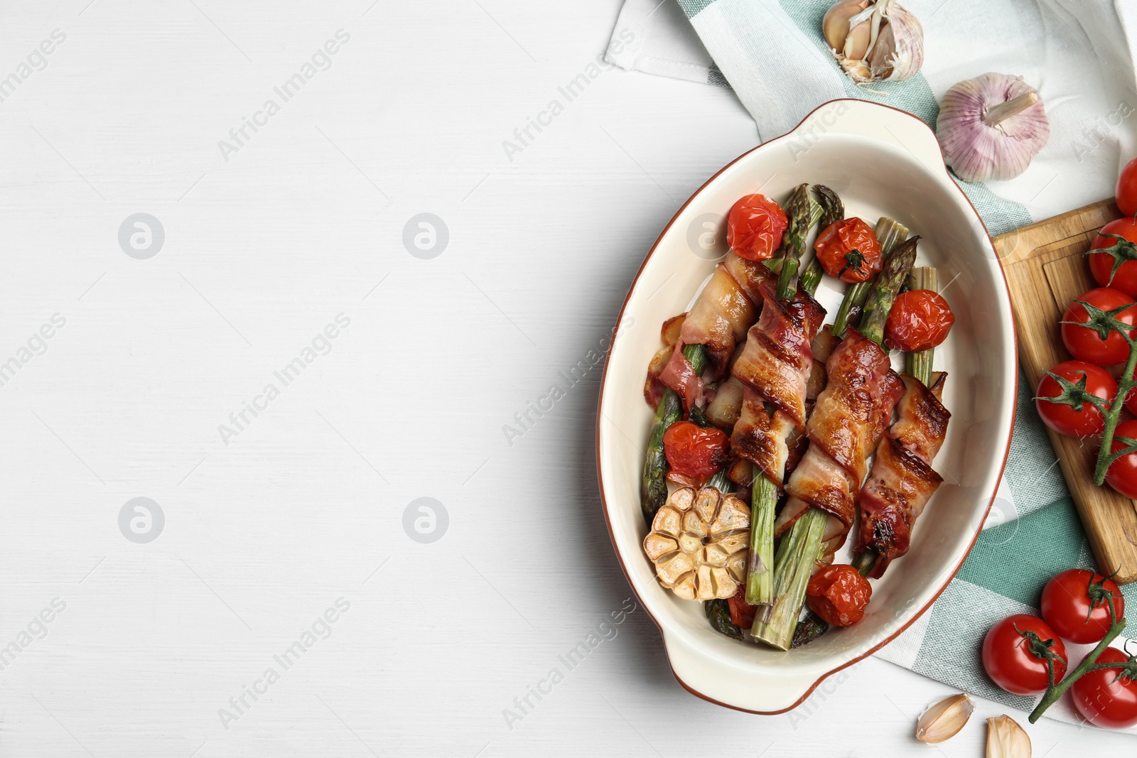 Photo of Oven baked asparagus wrapped with bacon in ceramic dish on white wooden table, flat lay. Space for text