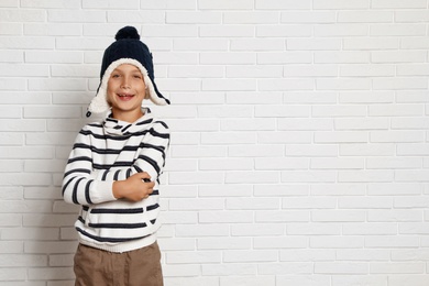 Photo of Cute little boy in hat and sweater near white brick wall, space for text. Winter season