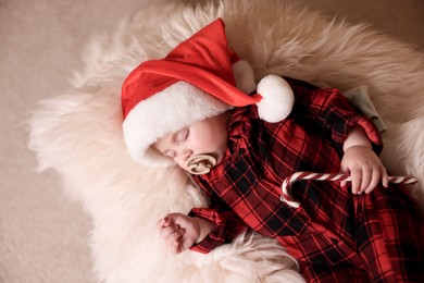 Cute baby in Santa hat with candy cane sleeping on soft faux fur, top view. Christmas season