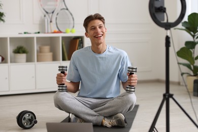 Photo of Smiling sports blogger holding dumbbells while streaming online fitness lesson at home