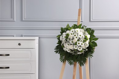 Photo of Funeral wreath of flowers on wooden stand near light grey wall indoors