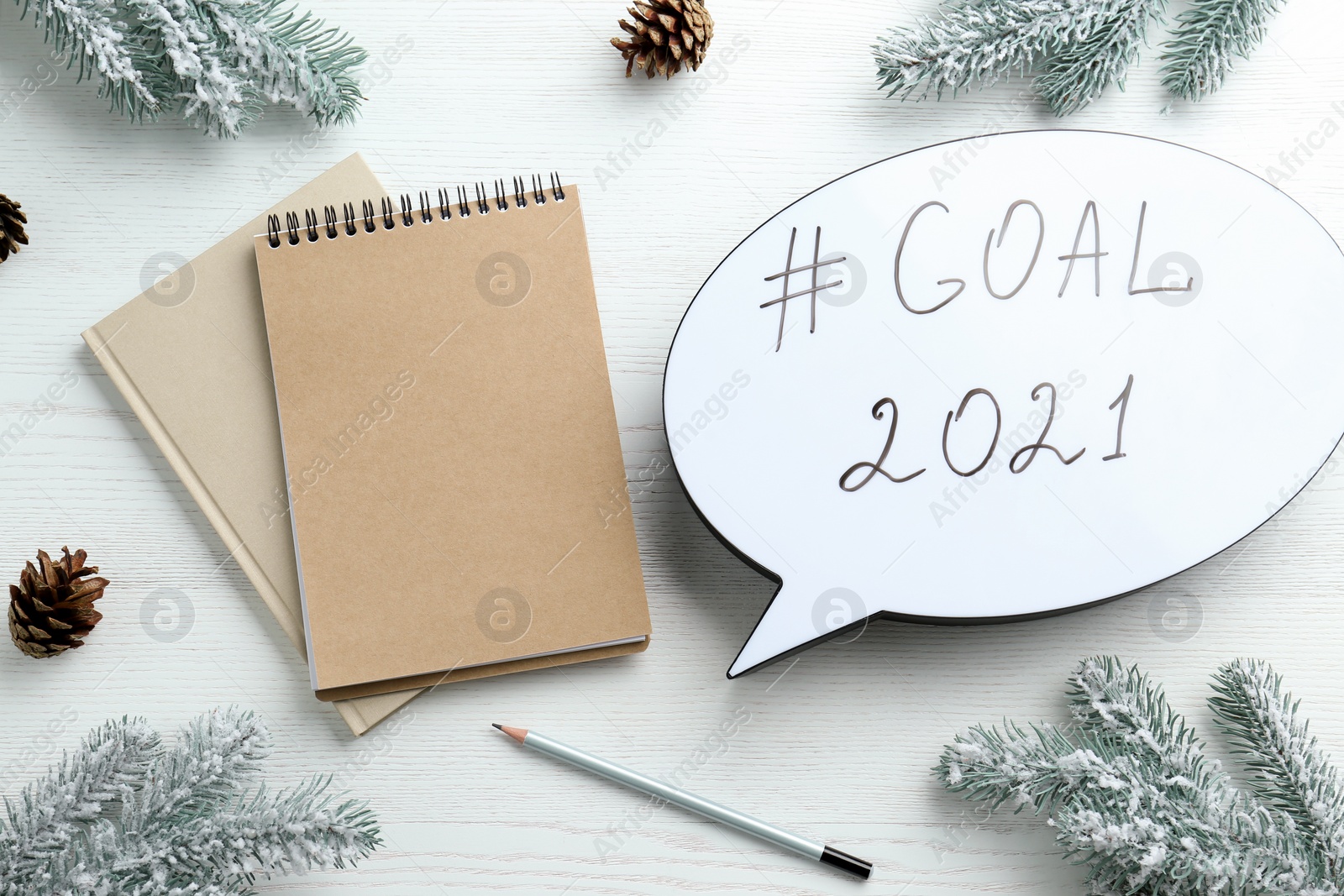 Photo of Paper speech bubble with hashtag Goal 2021, near notebooks and fir branches on white wooden table, flat lay. New year targets