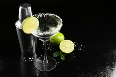 Photo of Metal shaker, delicious cocktail, limes and mint on black mirror surface, space for text